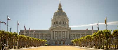 The grand, dome-topped San Francisco City Hall building viewed from a wide, sunlit plaza lined with trees.