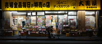 A lone shopper looks at goods outside a brightly lit discount shop in Tokyo at night.