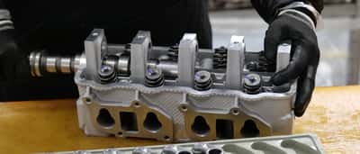 A worker wearing black gloves handles a newly machined metal engine component on a workbench.
