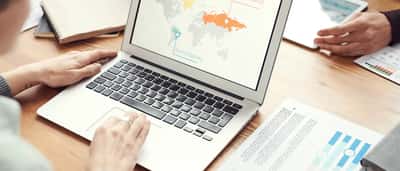 A person works on a laptop displaying a world map and data visualizations during a meeting at a wooden desk.