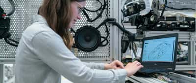 A female engineer reviews a 3D digital design on her laptop while working alongside testing equipment in an industrial workshop.