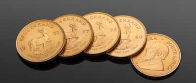 A row of gold Krugerrand coins resting on a dark, reflective surface.