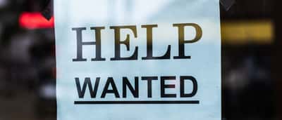 A red and white Help Wanted sign taped to the inside of a glass storefront window.