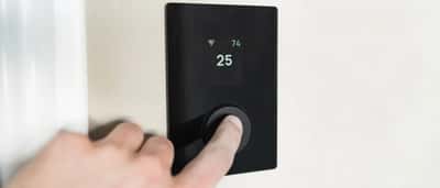 A close-up of a person's finger pressing the dial on a sleek, black digital smart thermostat mounted on a wall.