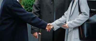 A man and a woman shake hands warmly across a table in a bright, modern office.