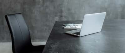 An empty black leather office chair sits next to a dark desk with an open laptop against a concrete wall.