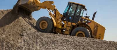 A yellow Caterpillar front loader carrying gravel up a steep mound under a clear blue sky.