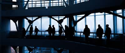 Silhouettes of people walking along the elevated walkways of a modern, multi-level glass office building.