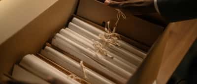 A professional in a grey suit carefully placing tied document scrolls into a cardboard archive box.