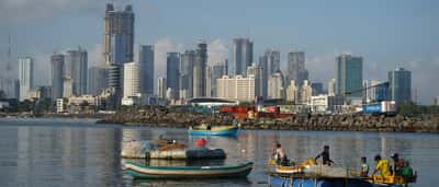 Traditional fishing boats float in a harbor with modern skyscrapers under construction in the background.