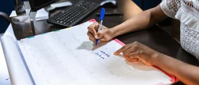 A professional writes notes on a large desk calendar in an office setting.