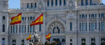 The iconic Palacio de Cibeles in Madrid adorned with waving Spanish flags under a bright blue sky.