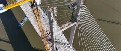 An aerial view of a yellow construction crane building a large cable-stayed bridge over a river.
