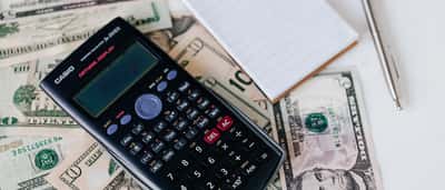 A black scientific calculator and a blank notepad sit on a scattered pile of US dollar bills.