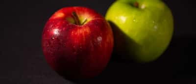 A red apple sits in sharp focus next to a blurred green apple against a dark background.