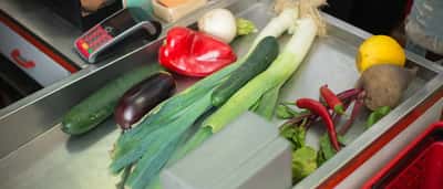 Fresh vegetables sit on a stainless steel grocery store checkout counter.