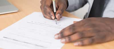 Close-up of a businessman in a grey shirt and tie signing a formal document with a fountain pen.