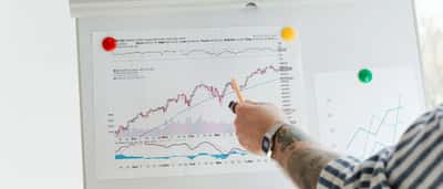 A person points with a pen to a printed stock chart of a broad U.S. market index pinned to a whiteboard.