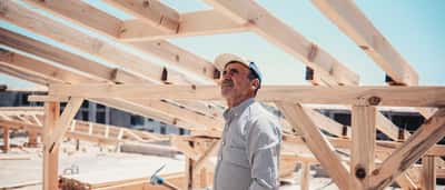 A construction worker holding a hammer stands beneath the exposed wooden roof trusses of a new home build.
