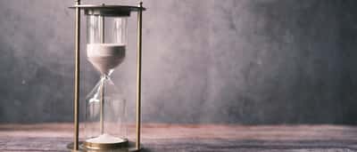 A brass hourglass filled with white sand sits on a wooden surface against a dark, mottled background.