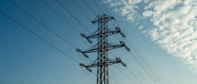 A tall electrical transmission tower stands against a bright blue sky with scattered clouds.