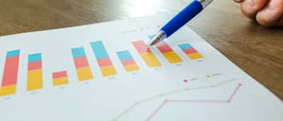 A person's hand holds a blue pen, pointing at a colorful printed financial bar chart resting on a wooden desk.