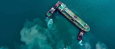 A top-down aerial view of an oil tanker being guided by two tugboats in deep turquoise waters.