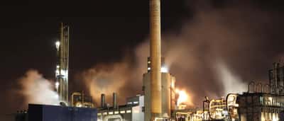 A sprawling industrial oil refinery illuminated at night, with smokestacks emitting thick plumes into the dark sky.