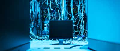 A dense, chaotic tangle of illuminated blue network cables surrounding a blank monitor in a dark server room.