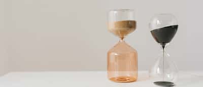 Two modern glass hourglasses filled with beige and black sand rest on a clean white desk.