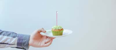 A hand in a striped button-down shirt holds a small plate with a green cupcake and a single lit candle.