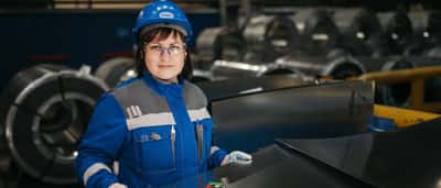 A woman in safety gear inspects large metal sheets in a modern industrial manufacturing setting.