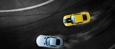 An aerial view of a yellow car and a white car drifting closely together around a curved asphalt track.