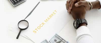 A person rests folded hands on a desk with stock market charts, a calculator, and scattered cash.