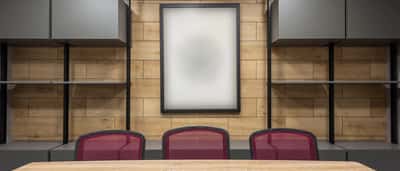 A blank white framed poster hangs on a wooden wall above a conference table and three red office chairs.