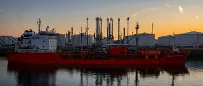 A large red oil tanker docked in front of an expansive oil refinery and storage tanks at dusk.