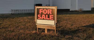 A real estate 'For Sale' sign stands in the front yard of a suburban home at sunset.
