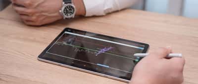 A person's hands hold a digital tablet displaying financial trading charts over a wooden desk.