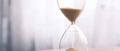 A clean, minimalist glass hourglass with sand flowing downward against a bright, soft background.