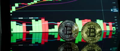 Two physical gold Bitcoin tokens stand on a reflective desk in front of a glowing monitor displaying a cryptocurrency trading chart.