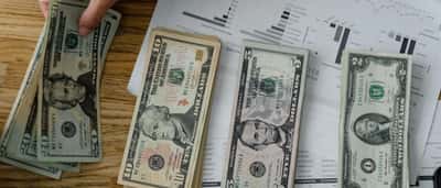 A person's hand counts US dollar bills over a desk covered in financial performance charts.