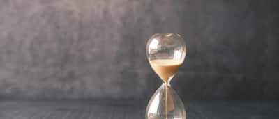 A close-up of a glass hourglass with white sand flowing downward, resting on a wooden surface.