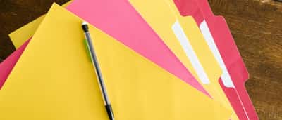A black pen rests on a neatly fanned stack of yellow and pink file folders on a wooden desk.