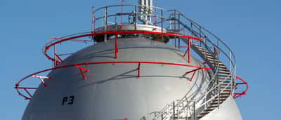A large, spherical industrial natural gas storage tank with a metal staircase winding around it against a blue sky.