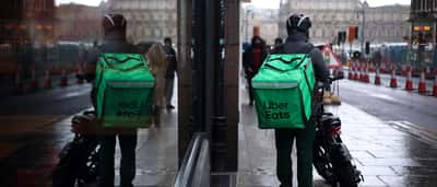 An Uber Eats courier wearing a green delivery backpack stands on a wet city street, reflected in a nearby window.