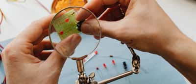 A person uses a magnifying glass to closely inspect a small electronic circuit board.