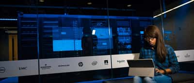 A woman works on a laptop in a server room behind a glass wall displaying the logos of various technology companies.