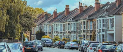 A row of classic British terraced houses stretches down a residential street beneath a partly cloudy sky.