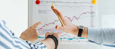 Two people pointing at a printed financial market chart pinned to a whiteboard.