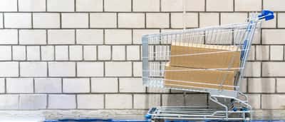 Plain cardboard boxes sit inside a metal shopping cart against a white brick wall.